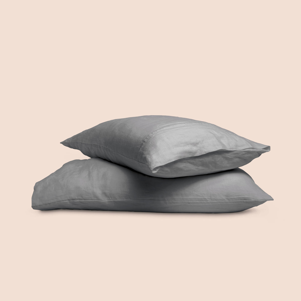 Image of two pillows with Stone Gray Relaxed Hemp pillowcases stacked on top of each other on a light pink background. The top pillow is showcasing an enveloping feature. 