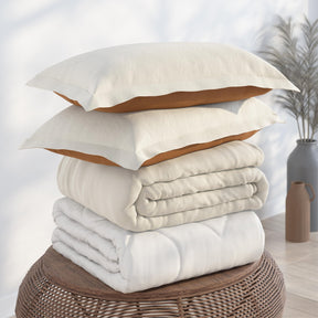 Image of a pile of bedding on top of a brown table. The bedding shown includes (from top to bottom): 2 Ivory/Clay Pillow Shams with the Ivory side facing up, a neatly folded Ivory/Clay Duvet Cover + Cooling with the ivory side showing, and a neatly folded Cooling Duvet Insert 