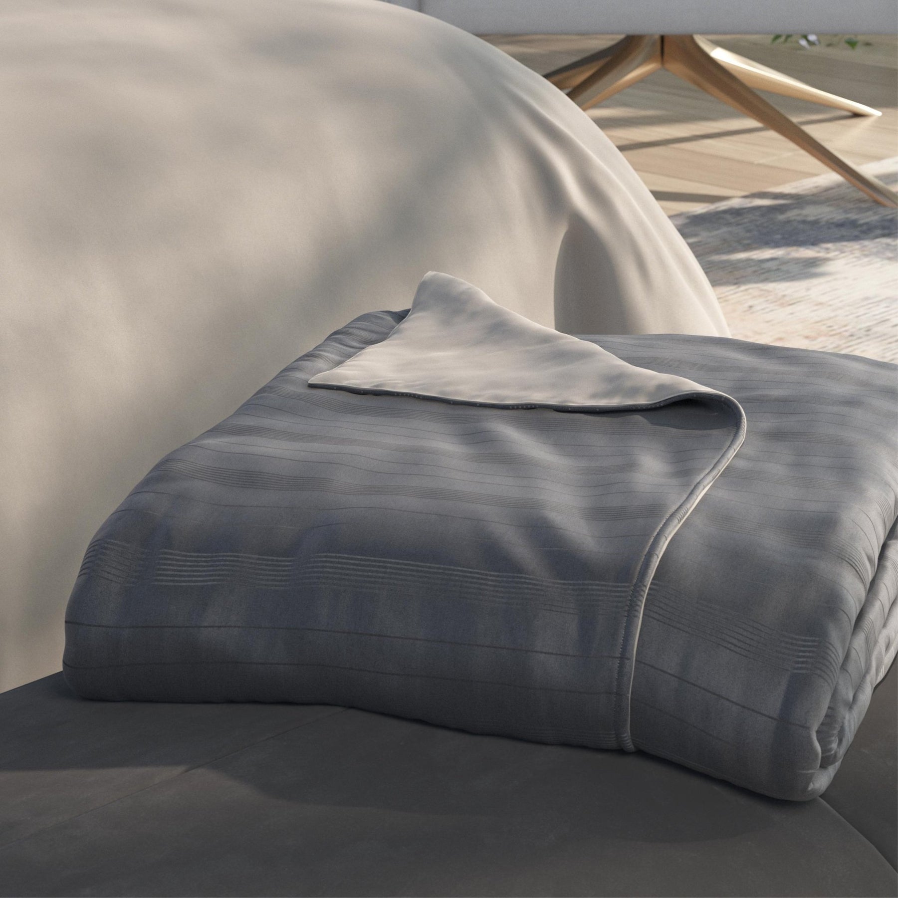 Image of a neatly folded Shadow/Dove Gray Duvet Cover + Cooling with the Shadow side facing up and a corner of the Dove Gray side folded back on a gray stool at the foot of a bed