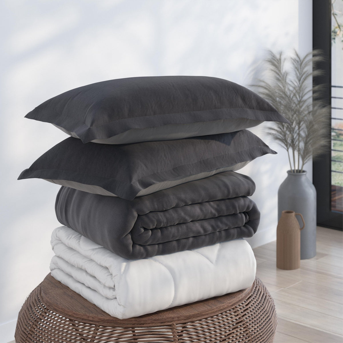 Image of a pile of bedding on top of a brown table. The bedding shown includes (from top to bottom): 2 Shadow/Dove Gray Pillow Shams with the Shadow side facing up, a neatly folded Shadow/Dove Gray Duvet Cover + Cooling with the Shadow side showing, and a neatly folded Cooling Duvet Insert 