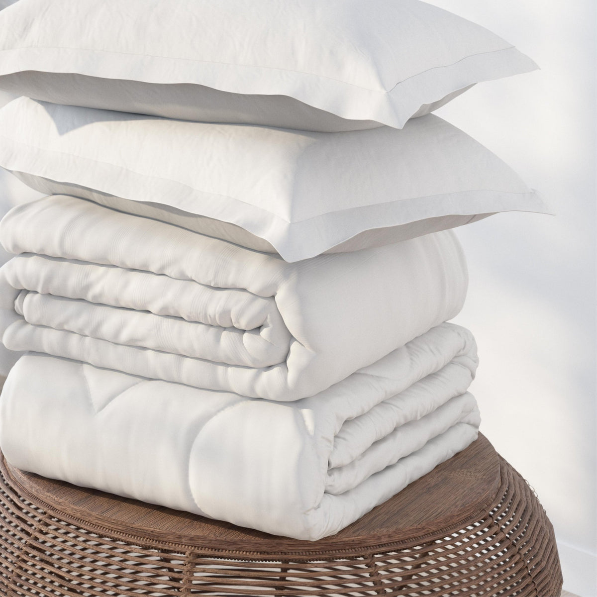 Image of a pile of bedding on top of a brown table. The bedding shown includes (from top to bottom): 2 White Pillow Shams, a neatly folded White Duvet Cover + Cooling, and a neatly folded Cooling Duvet Insert