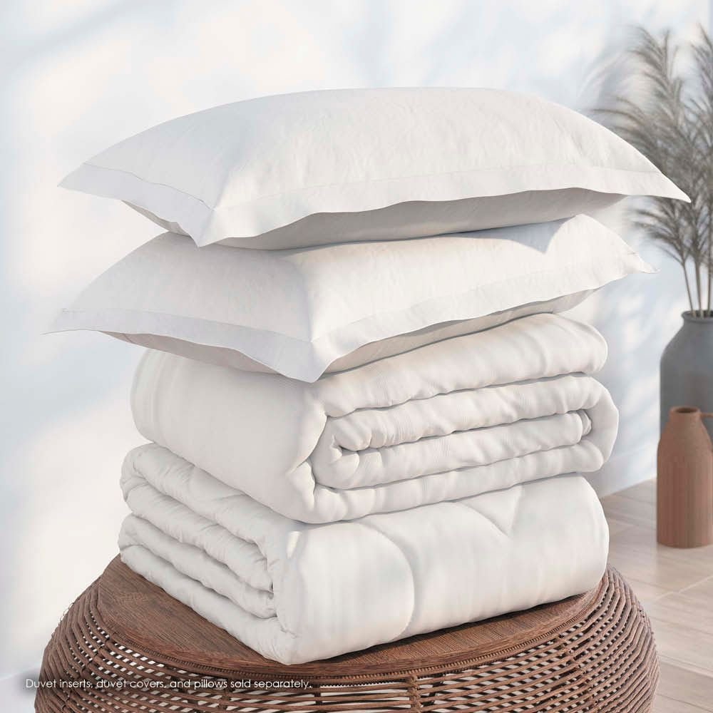 Image of a pile of bedding on top of a brown table. The bedding shown includes (from top to bottom): 2 White Pillow Shams, a neatly folded White Duvet Cover + Cooling, and a neatly folded Cooling Duvet Insert 