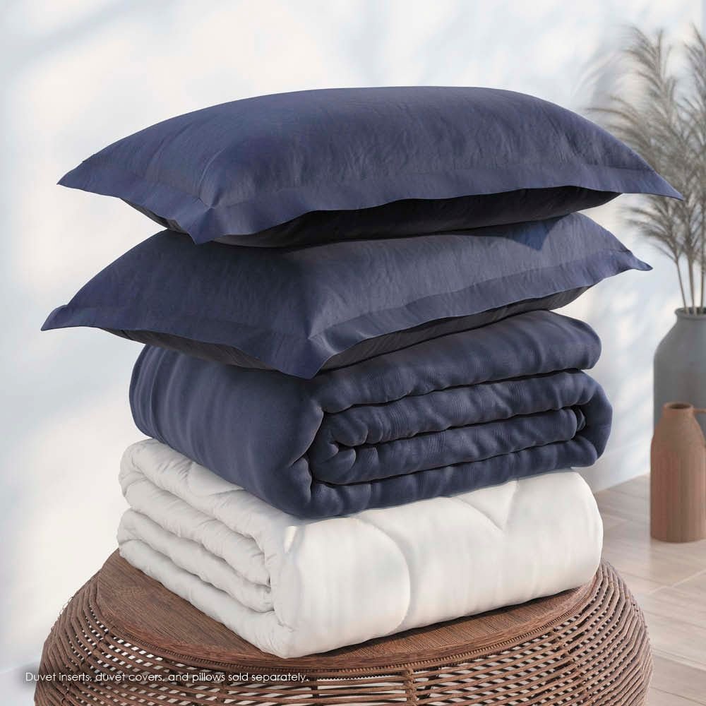 Image of a pile of bedding on top of a brown table. The bedding shown includes (from top to bottom): 2 Midnight/Celestial Pillow Shams with the Midnight side facing up, a neatly folded Midnight/Celestial Duvet Cover + Cooling with the Midnight side showing, and a neatly folded Cooling Duvet Insert 