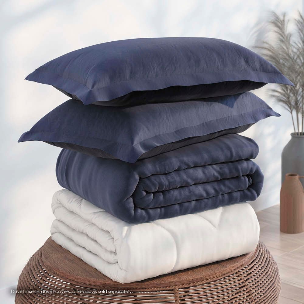 Image of a pile of bedding on top of a brown table. The bedding shown includes (from top to bottom): 2 Midnight/Celestial Pillow Shams with the Midnight side facing up, a neatly folded Midnight/Celestial Duvet Cover + Cooling with the Midnight side showing, and a neatly folded Cooling Duvet Insert