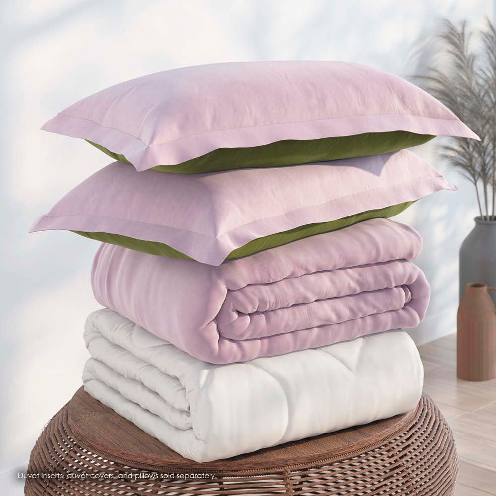 Image of a pile of bedding on top of a brown table. The bedding shown includes (from top to bottom): 2 Lilac/Jungle Pillow Shams with the Lilac side facing up, a neatly folded Lilac/Jungle Duvet Cover + Cooling with the Lilac side showing, and a neatly folded Cooling Duvet Insert 