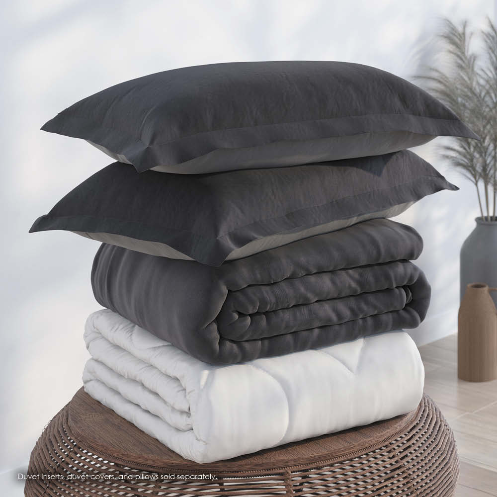 Image of a pile of bedding on top of a brown table. The bedding shown includes (from top to bottom): 2 Shadow/Dove Gray Pillow Shams with the Shadow side facing up, a neatly folded Shadow/Dove Gray Duvet Cover + Cooling with the Shadow side showing, and a neatly folded Cooling Duvet Insert