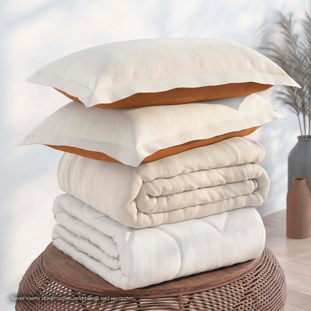 Image of a pile of bedding on top of a brown table. The bedding shown includes (from top to bottom): 2 Ivory/Clay Pillow Shams with the Ivory side facing up, a neatly folded Ivory/Clay Duvet Cover + Cooling with the Ivory side showing, and a neatly folded Cooling Duvet Insert 