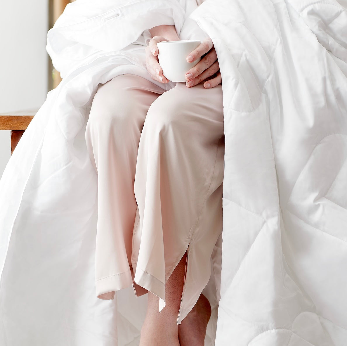 Image of a woman in light pink pajamas wrapped in a white duvet insert holding a cup of coffee. 