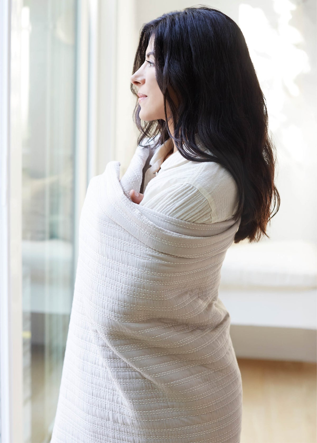 Image of a woman wrapped in a Bright White Everyday Cotton Coverlet staring out a wintery window