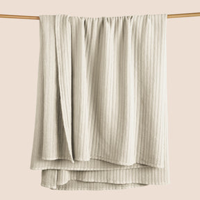 Image of an Ecru Ridgeback Coverlet hanging and draped over a wooden rod on a light pink background