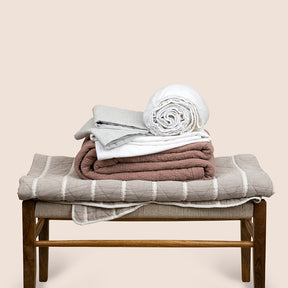 Image showcasing the Heritage Bundle, featuring the a brown bench with folded bedding on top. From bottom to top: gray and white Heritage Quilt, Pink Sandstone Wave Coverlet, White Blended Linen Sheets, Pinstripe Relaxed Hemp Pillowcases.