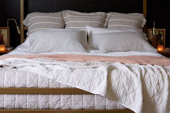 Image of a completely dressed bed with dark background. Bed is dressed with white sheets, two pillows with gray pillowcases, two pillows with white pillowcases, three Euro Pillows with Heritage Pillow Shams, a Pink Sandstone Wave Coverlet, and a reversible white and gray Heritage Quilt.
