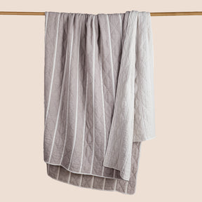 Image of a gray and white striped Heritage Quilt hanging and draped over a wooden rod on a light pink background