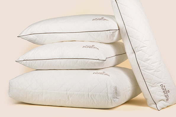 Image of a pile of Dr. Weil + Purecare pillows on a light pink background. Two Euro Pillow Inserts are stacked on top of one Meditation Cushion with a Lumbar Pillow propped up next to them.
