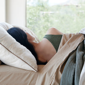 Image of a woman asleep in bed with her back facing the camera. The bed has Ochre Garment Washed Percale Sheets with an Agave Ridgeback coverlet over it. 