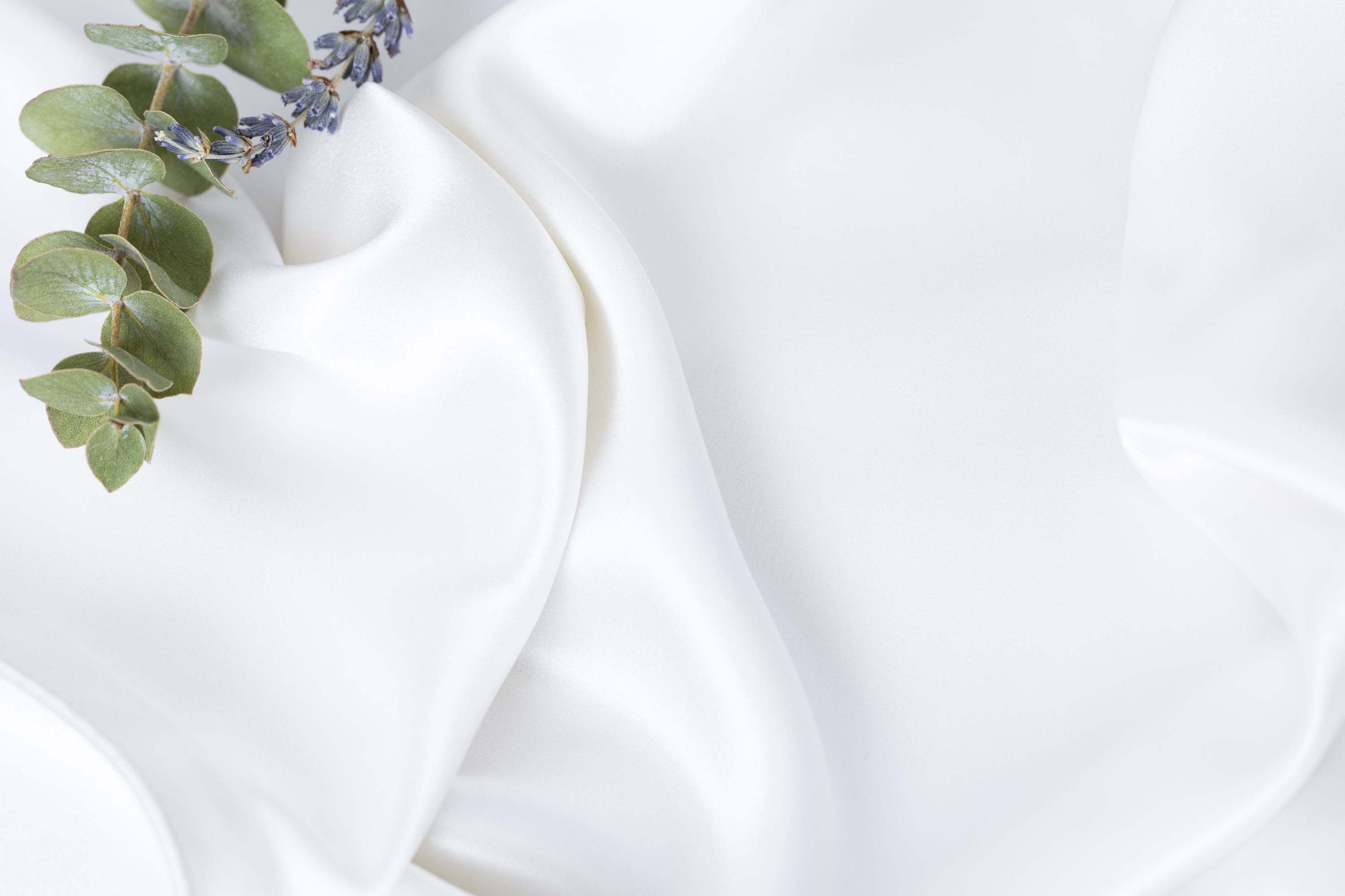 Close-up image of white silky fabric with lavender and eucalyptus sprigs in the top left corner