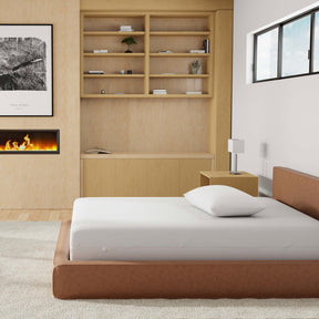 Image of a brown leather bed with just a mattress protector and pillow with pillow protector on the bed. The bed is in a light, neutral-colored room with a fireplace, picture of wood on the log, and wooden bookshelves.