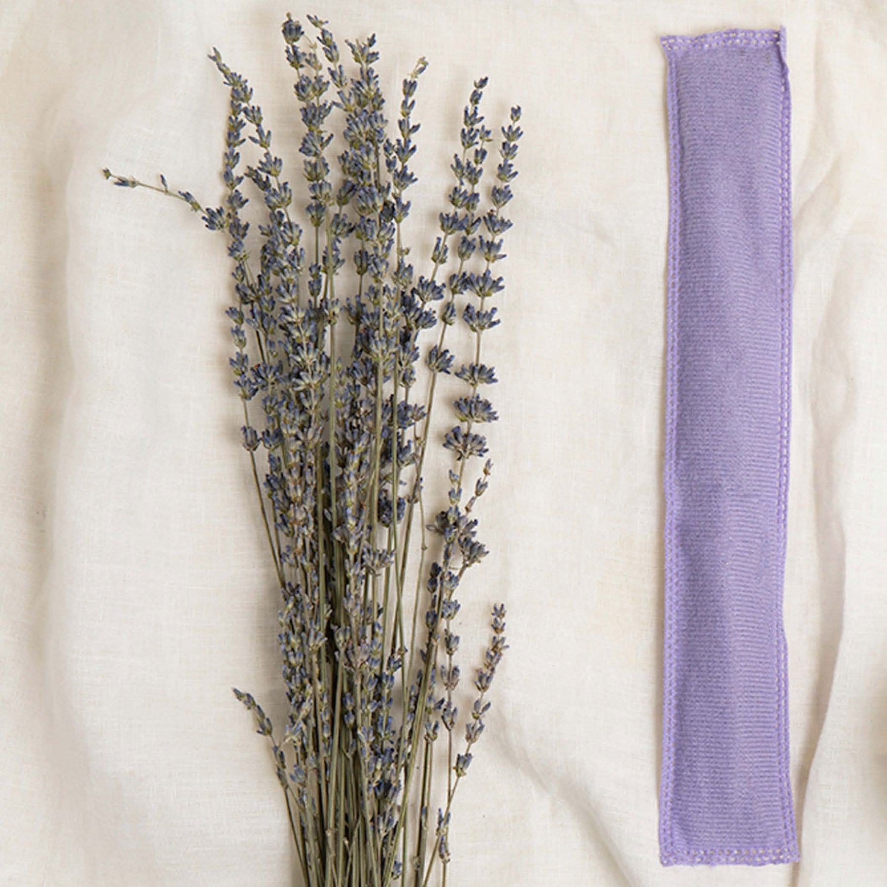 Image of a purple Aromatherapy sachet with lavender sprigs to the left of it on an off-white fabric background
