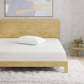 Image of a wooden bed with just a mattress protector and pillow with pillow protector on the bed. The bed is in a light, neutral-colored room with a purple painting above the bed and purple candles on the nightstand to the right of the bed. 