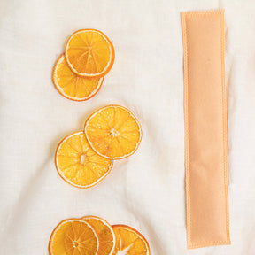 Image of an orange Aromatherapy sachet with 7 slices of mandarin oranges to the left of it on an off-white fabric background