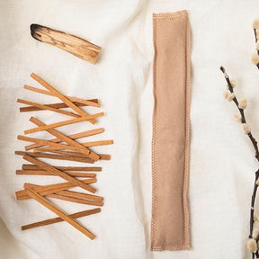 Image of a light tan Aromatherapy sachet with various pieces of wood to the left of it and a cotton sprig to the right on an off-white fabric background