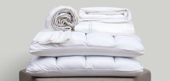 Image of a pile of all-white bedding on a taupe colored stool. The bedding includes (from top to bottom): a rolled white Refreshing TENCEL™ Lyocell fitted sheet, two white Refreshing TENCEL™ Lyocell Pillowcases, a neatly folded Cooling Mattress Protector, a neatly folded Refreshing TENCEL™ Lyocell flat sheet, and two SoftCell® Chill Pillows
