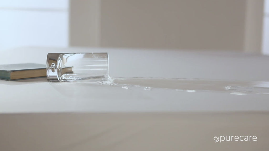 Video of water spilling on a mattress protector. A woman comes and takes the protector off of her bed and puts in her washer. She then remakes the bed and highlights the thick dual-sided elastic band on the bottom. 