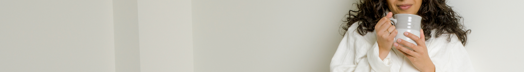 Image of a woman leaning against a wall holding a cup of coffee by her face with the white Dr. Weil Featherweight Robe on