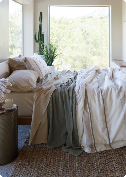 Side-view image of a made bed with a large window behind it. The bed showcases Ochre Garment Washed Percale Sheets, an Agave Ridgeback Coverlet, and a Sonoran Duvet Cover.