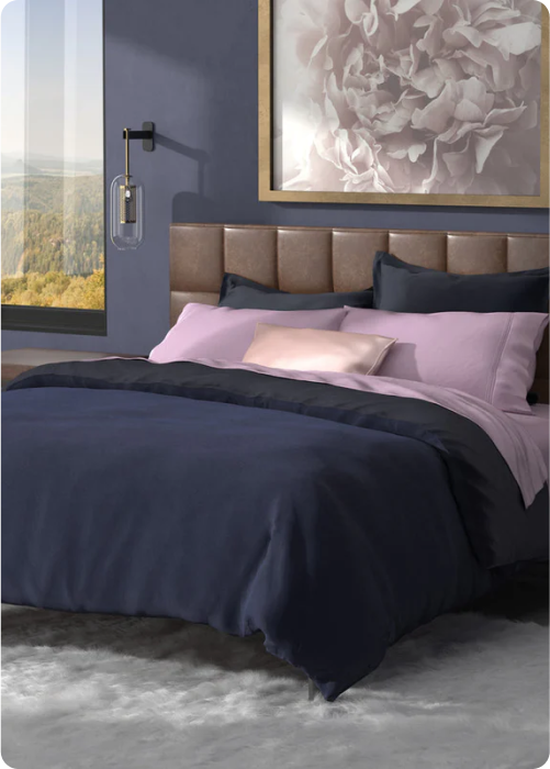 Image of a neatly made bed with Midnight/Celestial Blue Duvet Cover and Pillow Shams, Lilac sheets, and a Champaign Silk Pillowcase in a navy-colored room