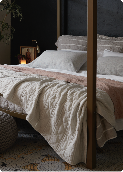 Image of a made bed with a dark background. The bed showcases pillows of various sizes and layered bedding. Items on the bed include: Kapok Pillows, White Blended Linen Sheets, Pinstripe Relaxed Hemp Pillowcases, a Pink Sandstone Wave Coverlet, and a reversible gray and white striped Heritage Quilt.