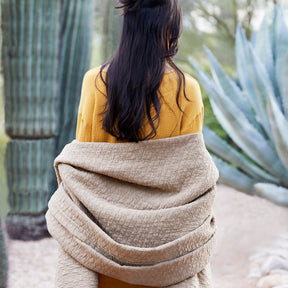 Image of a woman with long black hair and a yellow shirt standing outside by cacti. She is facing away, wrapped up in an Ochre Wave Coverlet. 