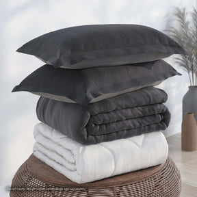 Image of a pile of bedding on top of a brown table. The bedding shown includes (from top to bottom): 2 Shadow/Dove Gray Pillow Shams with the Shadow side facing up, a neatly folded Shadow/Dove Gray Duvet Cover + Cooling with the Shadow side showing, and a neatly folded Cooling Duvet Insert 